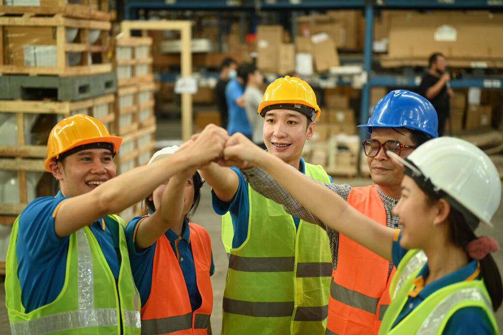 Diverse group of youth participating in a program funded by the Skills Development Program. All youth are wearing multicoloured hardhats in a warehouse setting, and they are raising their fists in celebration.